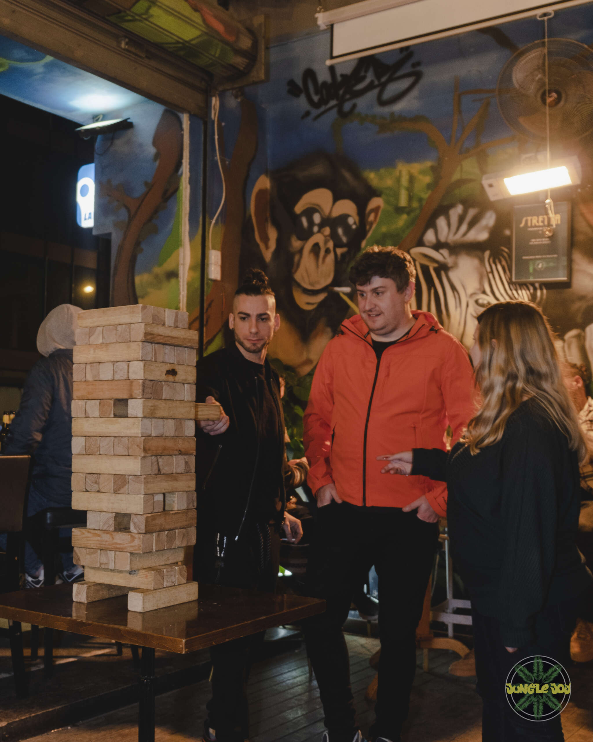 Three Players Enjoying Giant Jenga, One Adding a Piece, with Jungle Joy Mural Enhancing the Atmosphere