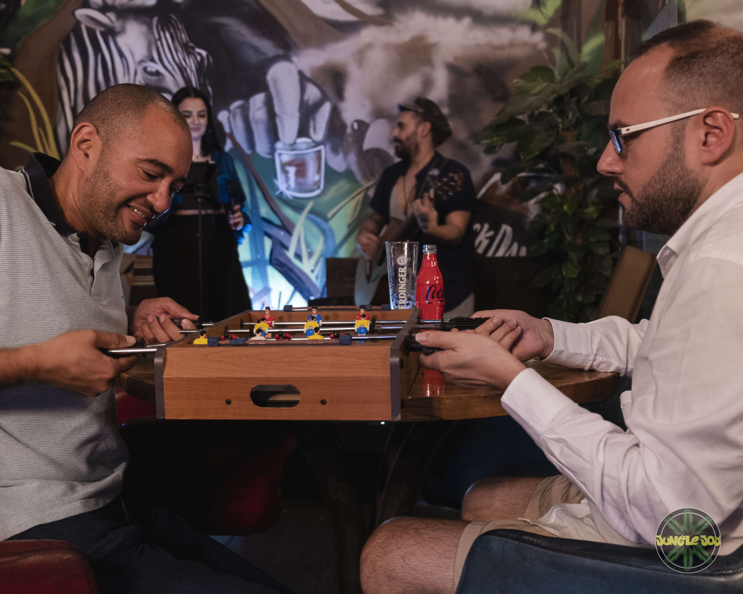 Two individuals engaged in a foosball game as a woman sings in the background.