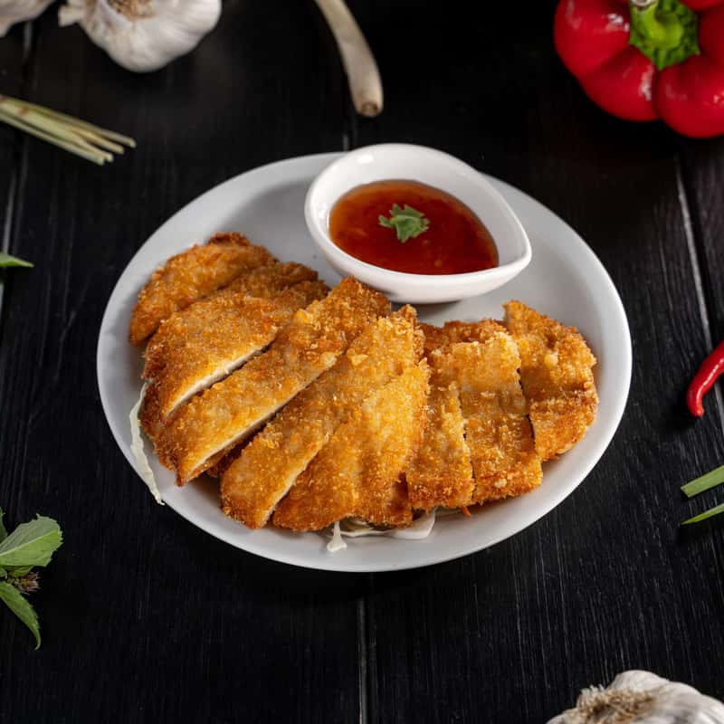 Marinated chicken breast with breadcrumbs and deep fried with sweet and sour sauce on a white plate, on a black table.